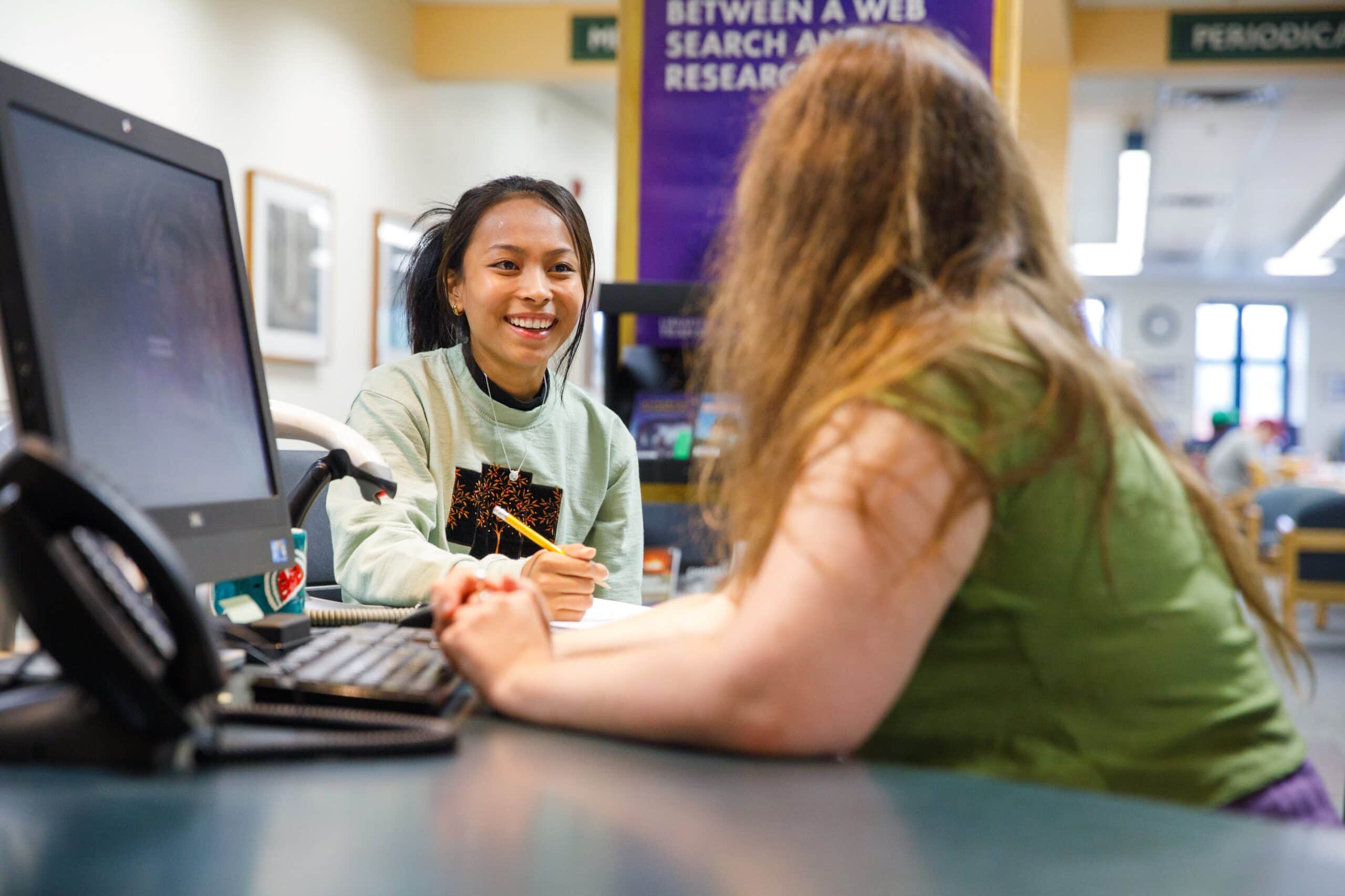 A student smiling at a librarian as they do research at a computer.