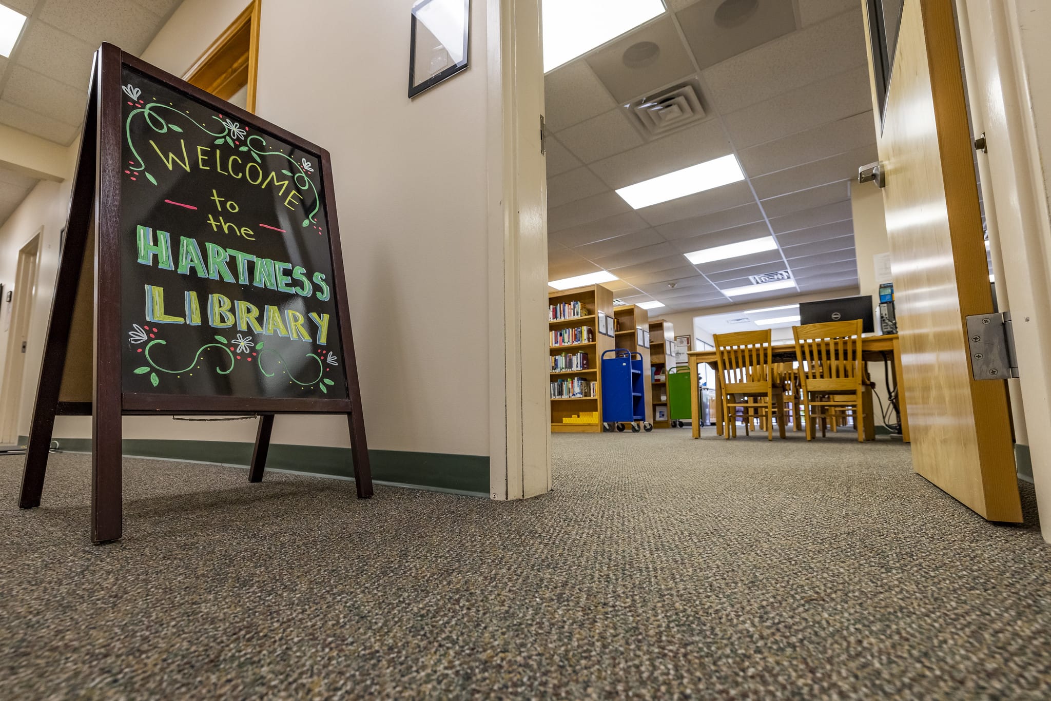 A sandwich board that says Hartness Library in a hallway next to a door that leads into a library.