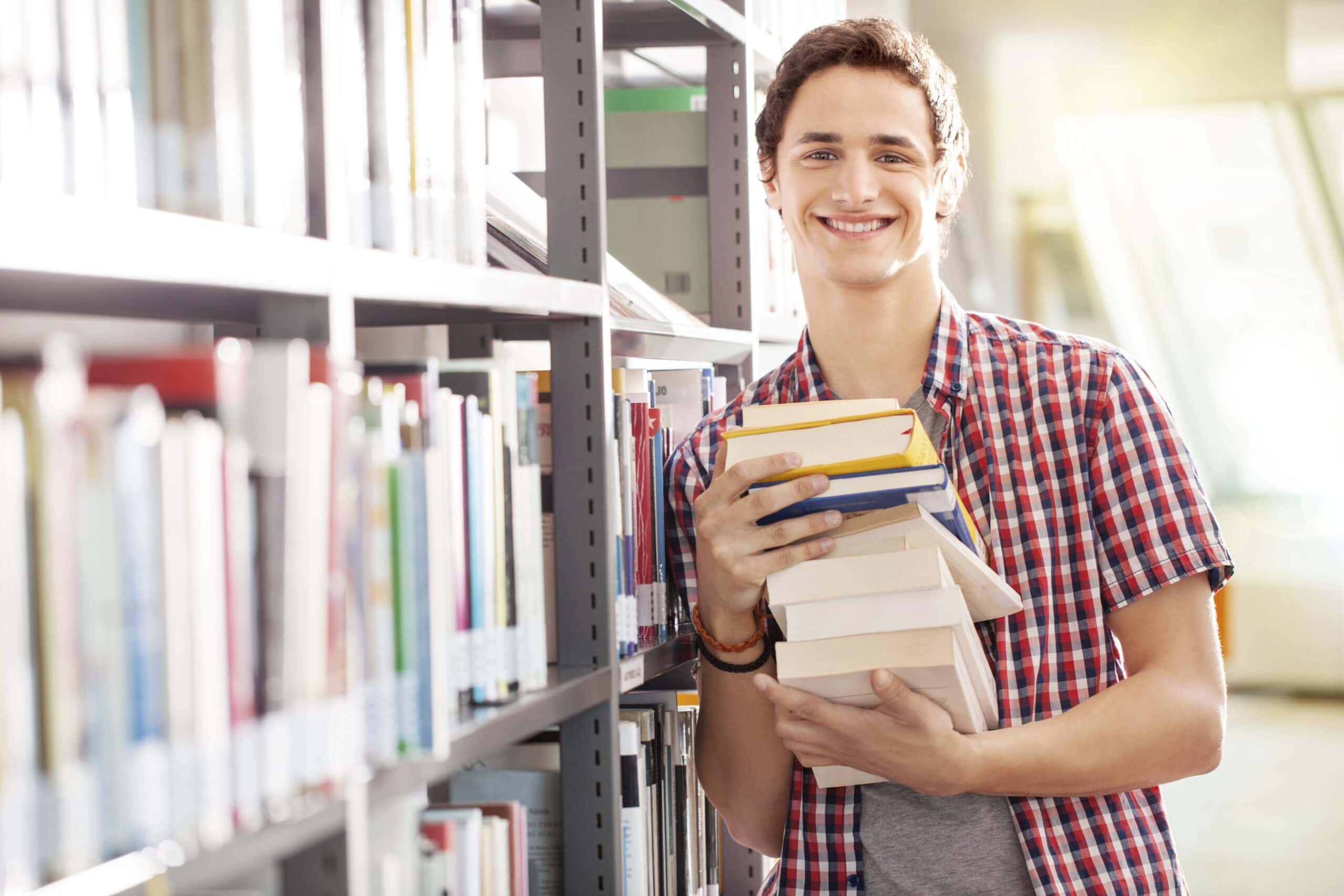 Portrait of a young student holding books standing by bookshelf in a library.