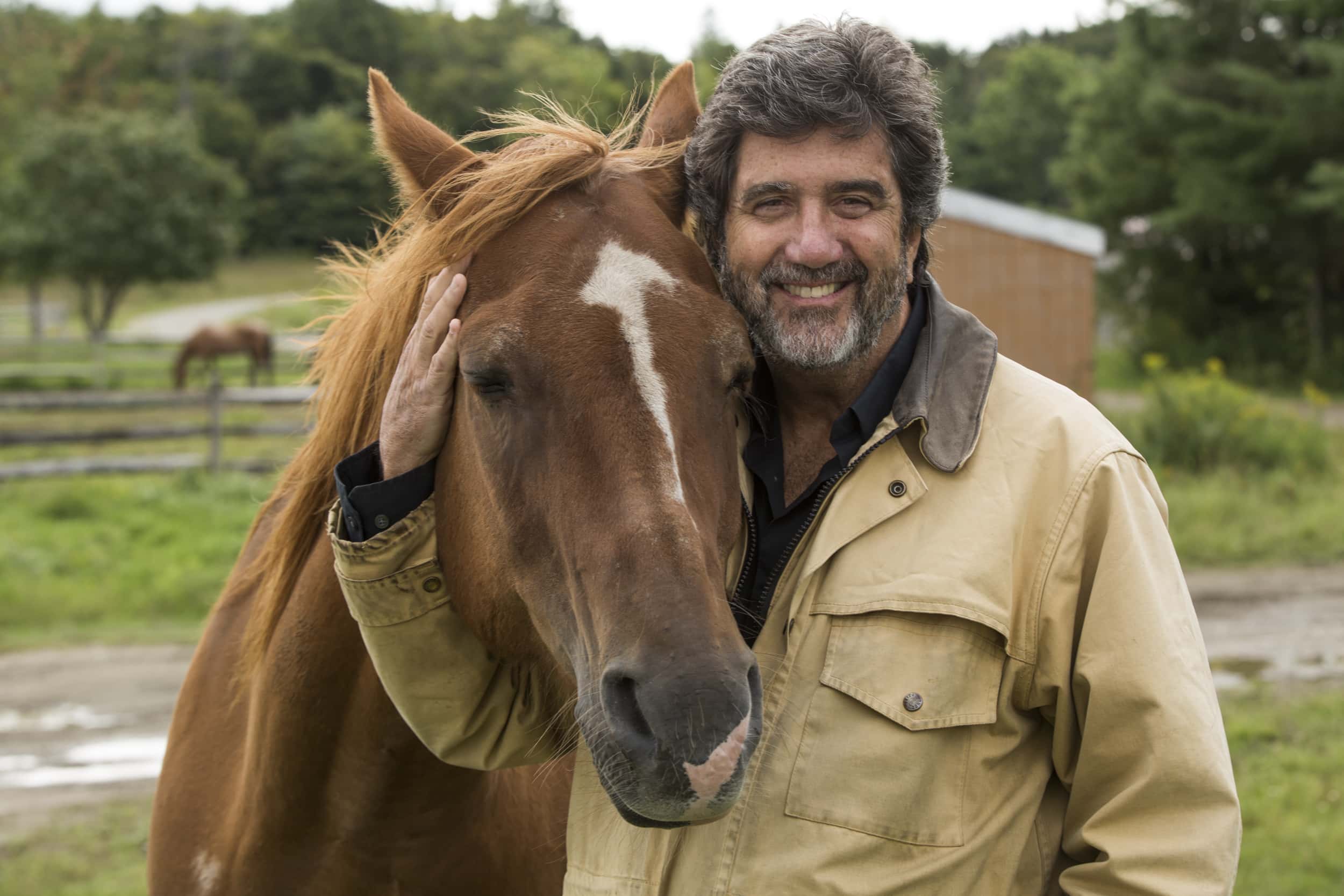 Timothy Hayer smiles at the camera with a arm wrapped around a light brown horse. With a field and another horse in the background.