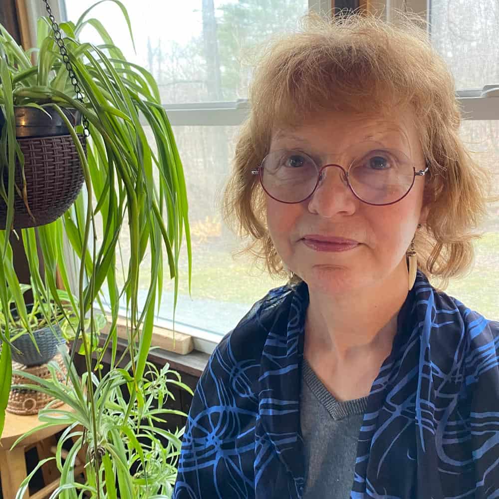 Woman in blue and black striped pattern jacket and glasses smiles at the camera with hanging plants in the background.