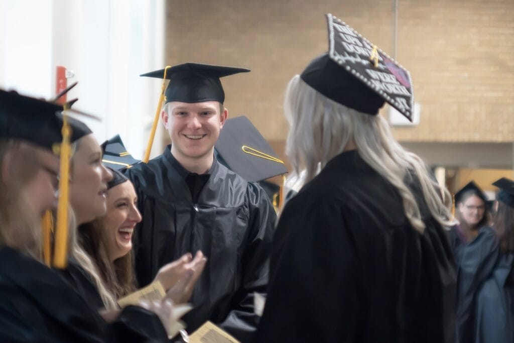 A young man in a cap and gown smiles in the center of a crowd of fellow Vermont State graduates.