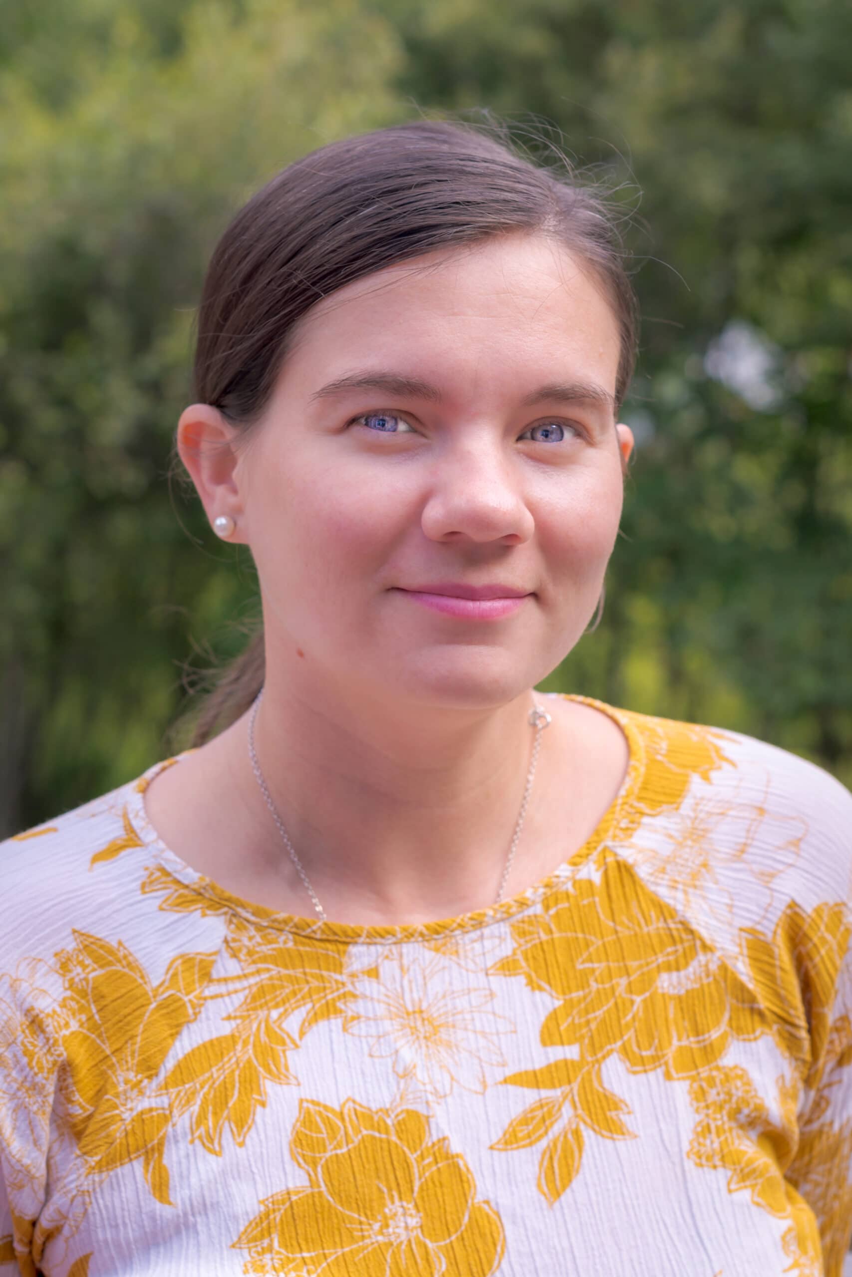 Sarah Truckle smiles at the camera in a yellow floral shirt and a forest in the background.