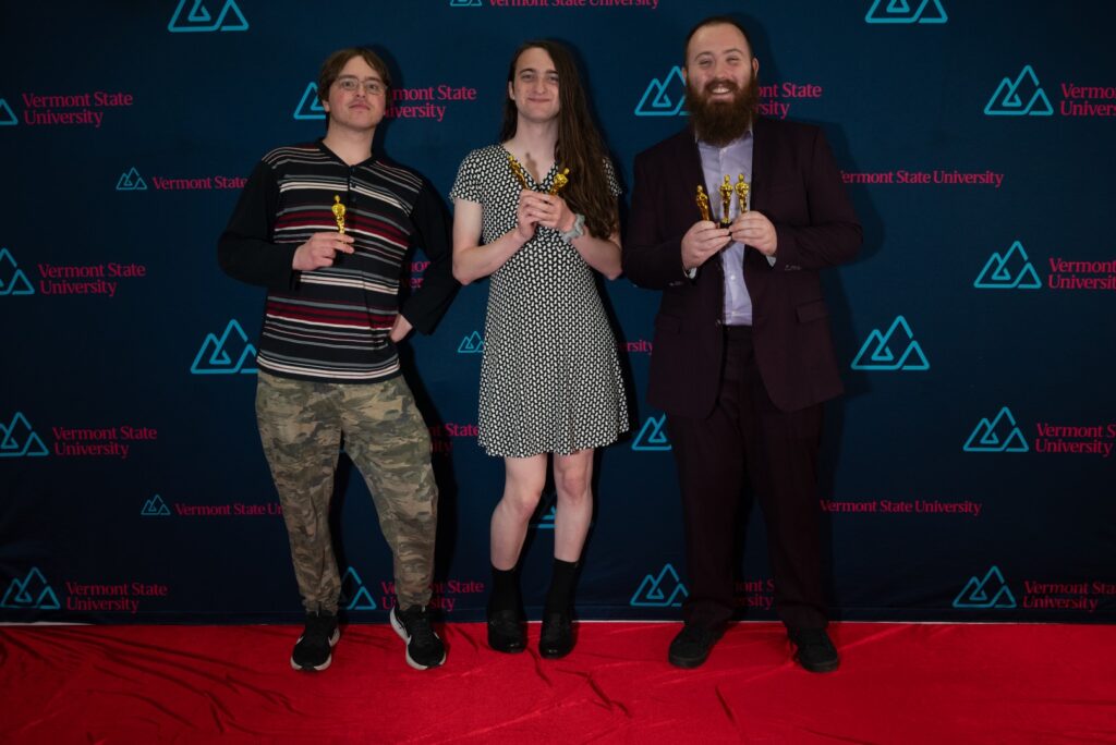 Three Vermont State University Students pose with golden statues on a red carpet.