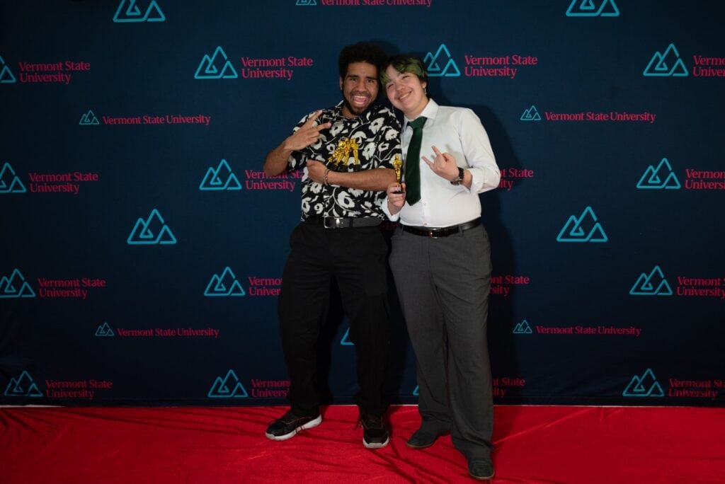 Two Vermont State University students embrace while holding golden statues on a red carpet.