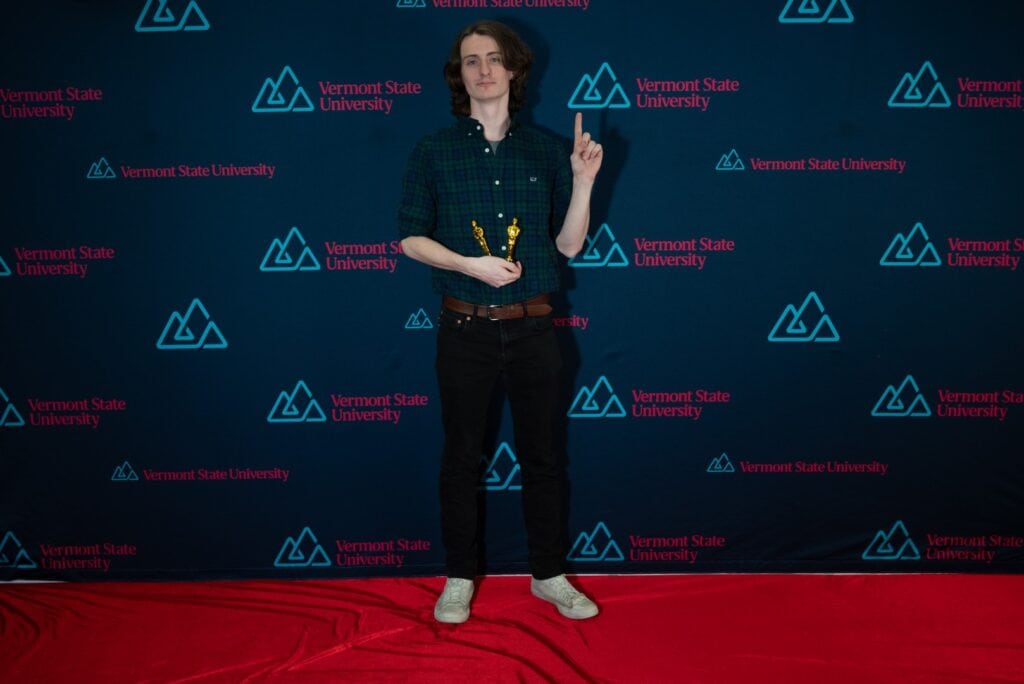 A young man dressed all in black with long hair points up while standing on a red carpet.