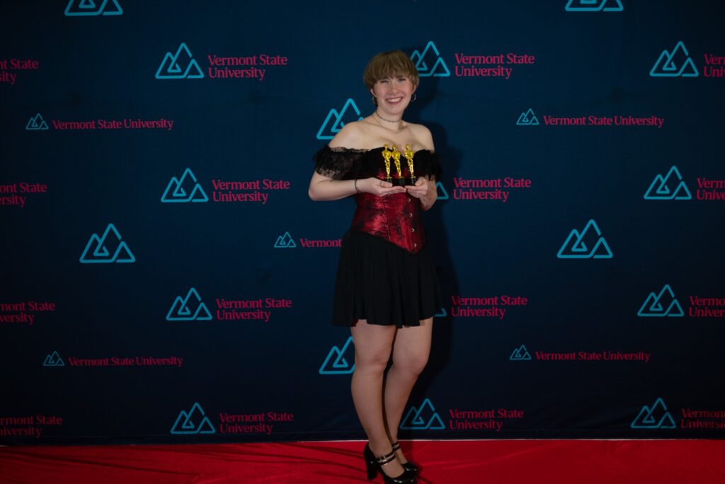 A young woman in a black dress holds a cluster of golden statues on a red carpet.