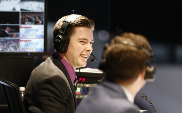 A young man wearing a headset and a suit smiles brightly at a colleague sitting next to him.