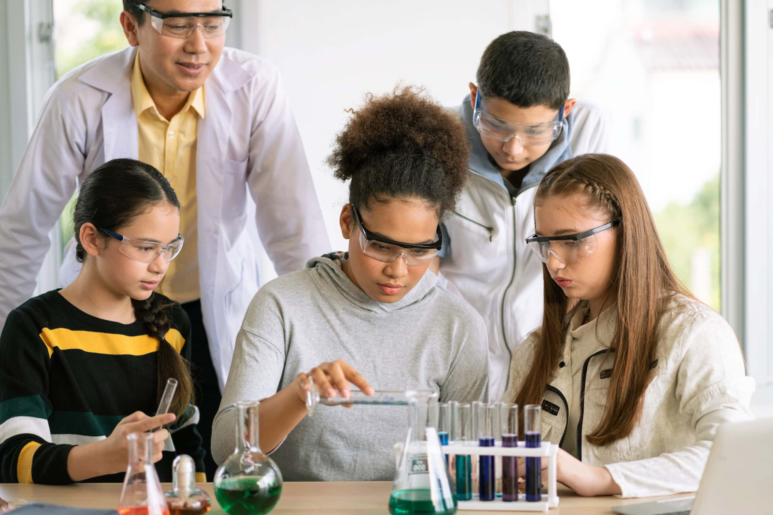 Diverse group of students in protective eyeglasses in science class doing chemical experiment in laboratory. Girl mixing chemistry in test tube.