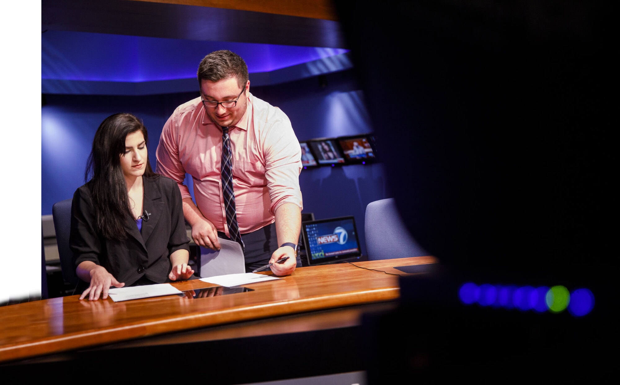 one person stands at a news desk as another sits and reads a script.