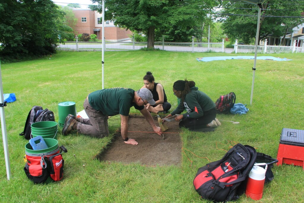 Three people hover over a rectangular hole in the ground taking earth samples.