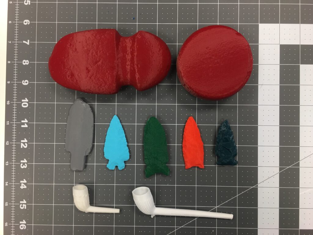 3D-printed copies of local artifacts used in educational outreach. A head of an axe 3d- printed red. 5 Arrow heads, each in a different color. Two white pipes sit below the Arrow heads.  