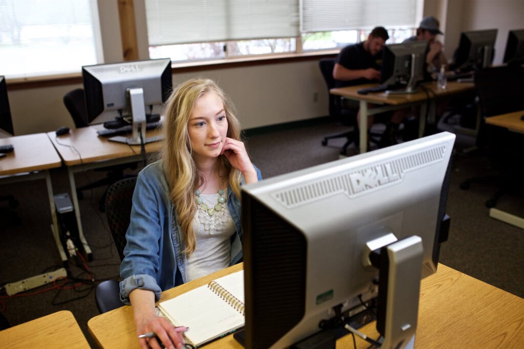 Person sits in a computer lab and is using a computer in a jean shirt and blonde hair.