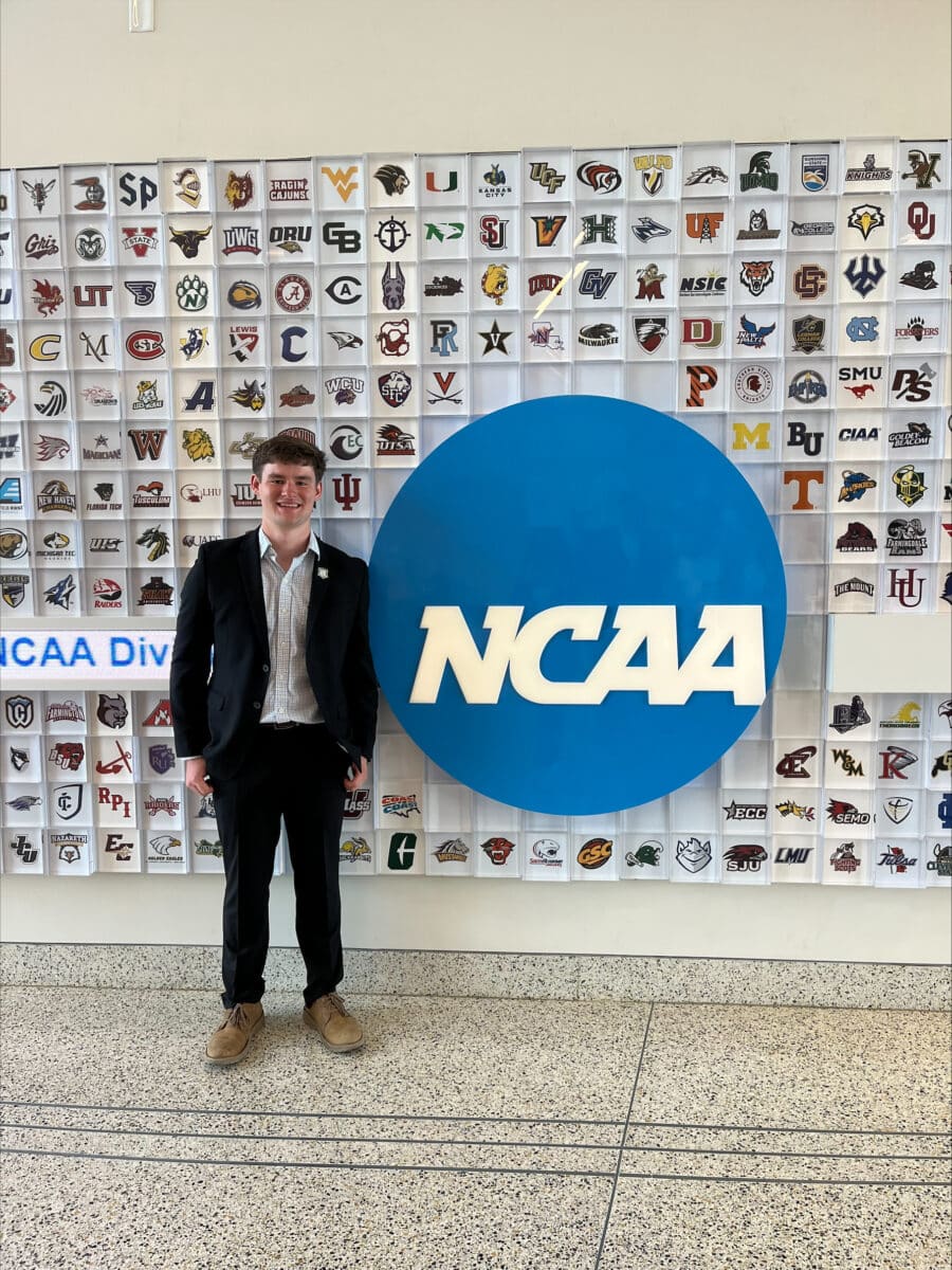 A young man in a suit stands in front of wall with the NCAA logo.