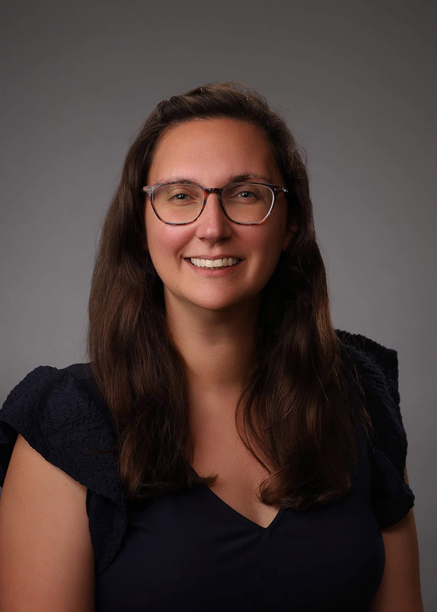 A woman with long brown hair and round glasses smiling at the camera.