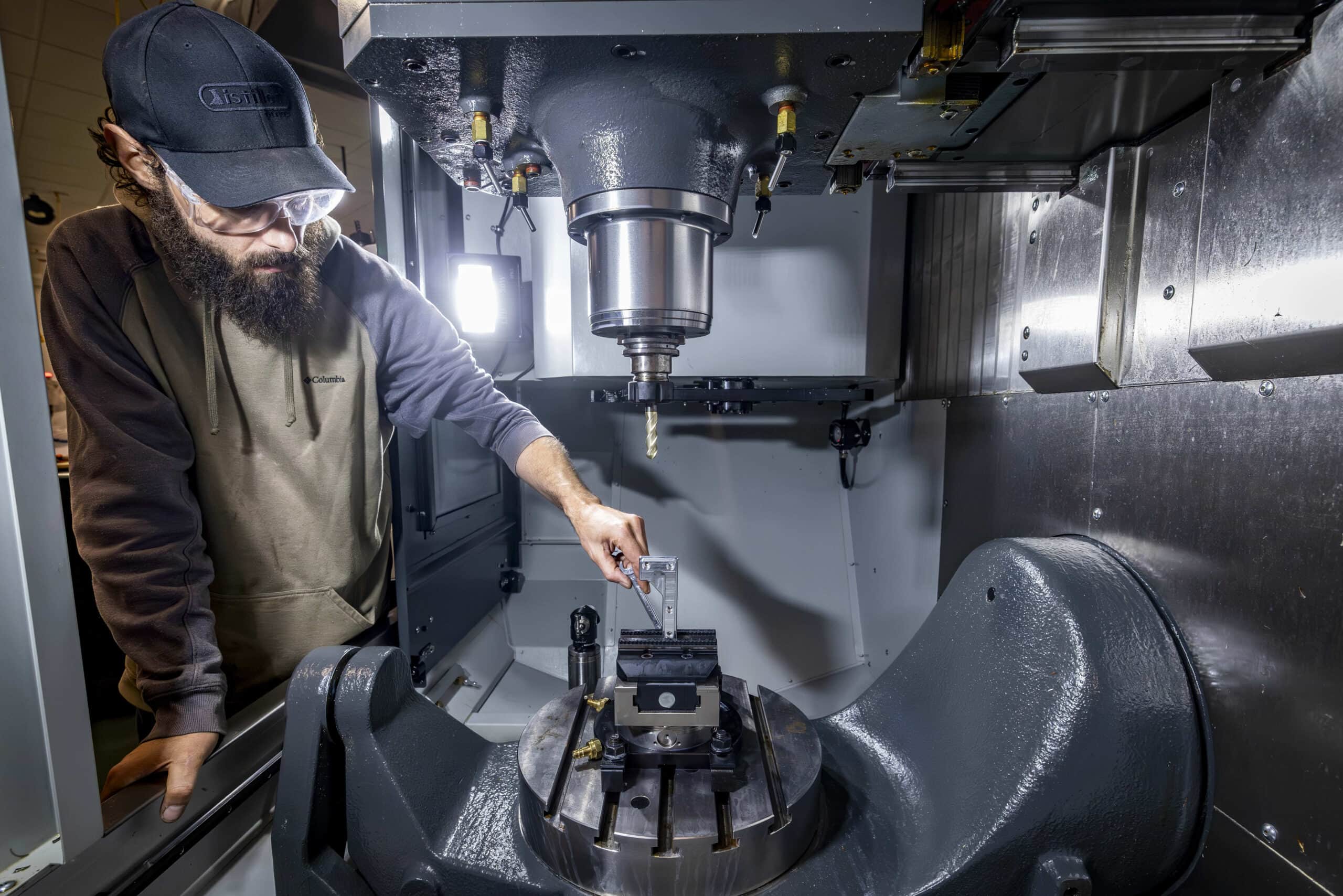 A man with a beard and a black baseball cap reaches his hand into a lathe.