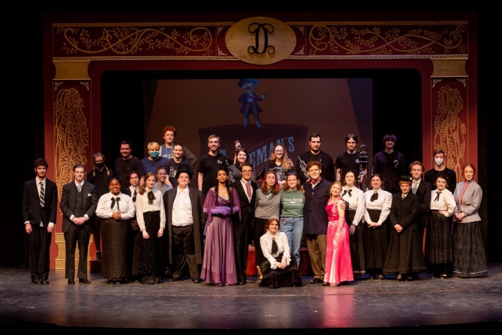 A large group of people in period garb stand and pose for a camera on the stage.