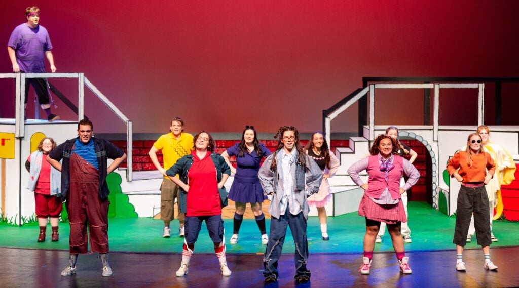 A group of people in colorful outfits stand with their hands on their hips on a stage.