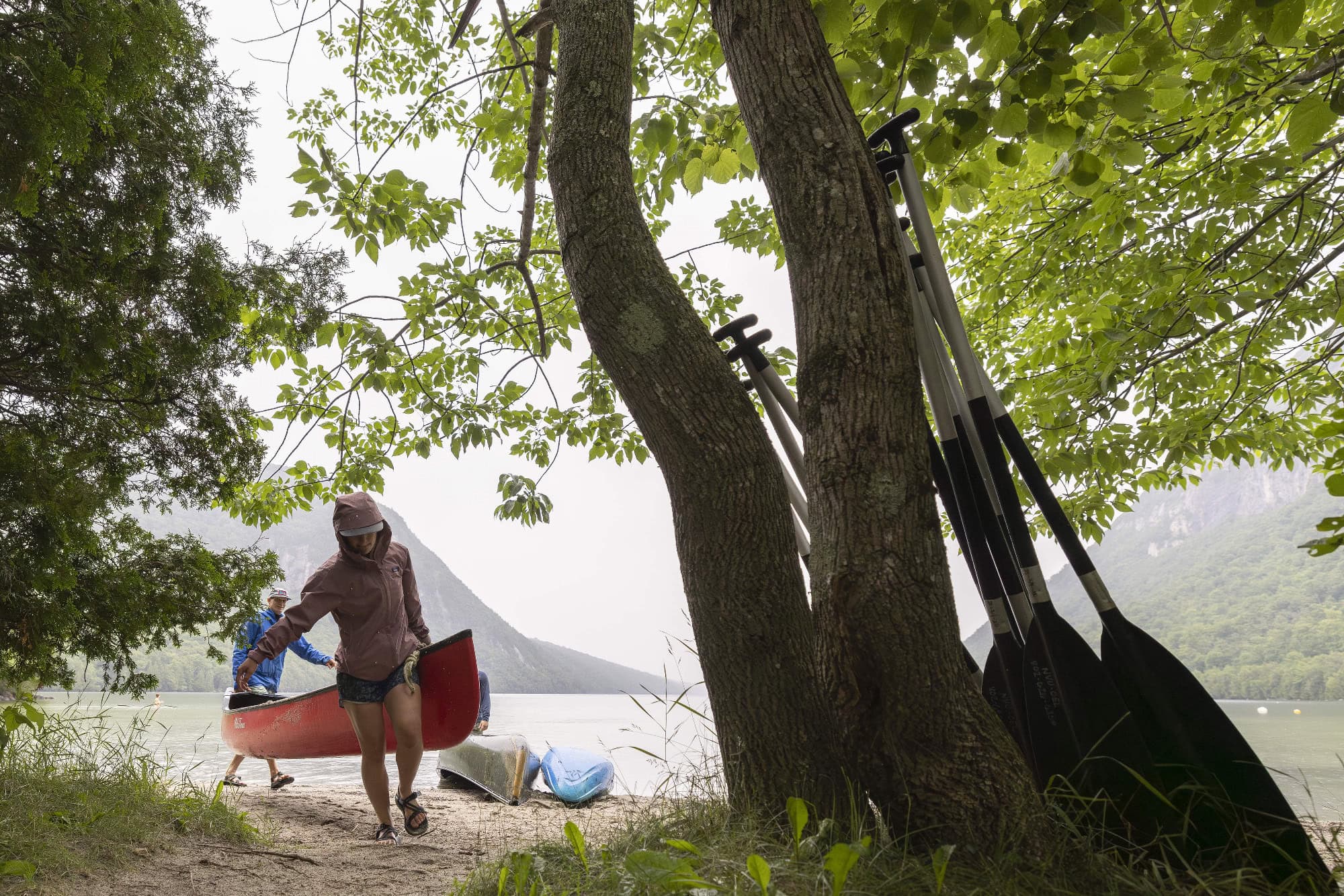 Two people carrying a red canoe along a wooded lakeshore, with paddles leaning against a tree and misty mountains in the background, capturing a serene outdoor adventure scene.