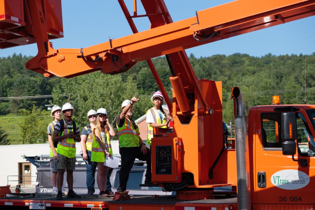 A volunteer from the transportation agency and students all wearing high-visibility vests stand and discuss a piece of orange equipment