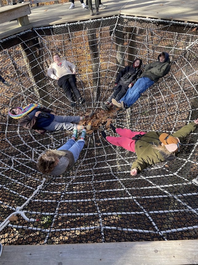 People lay in a circle on a spiderweb-like grid of netting with their feet all touching in the center.