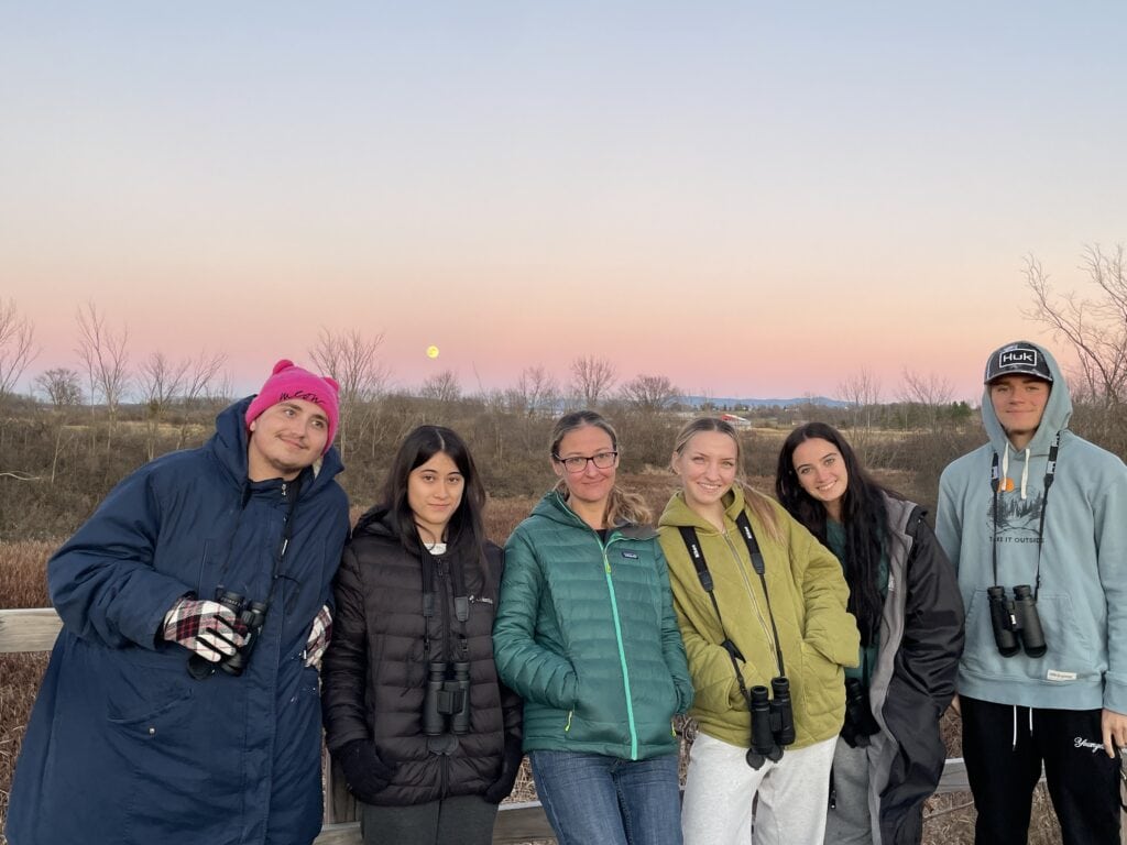 Students smile in front of a sunset, some have binoculars around their necks.