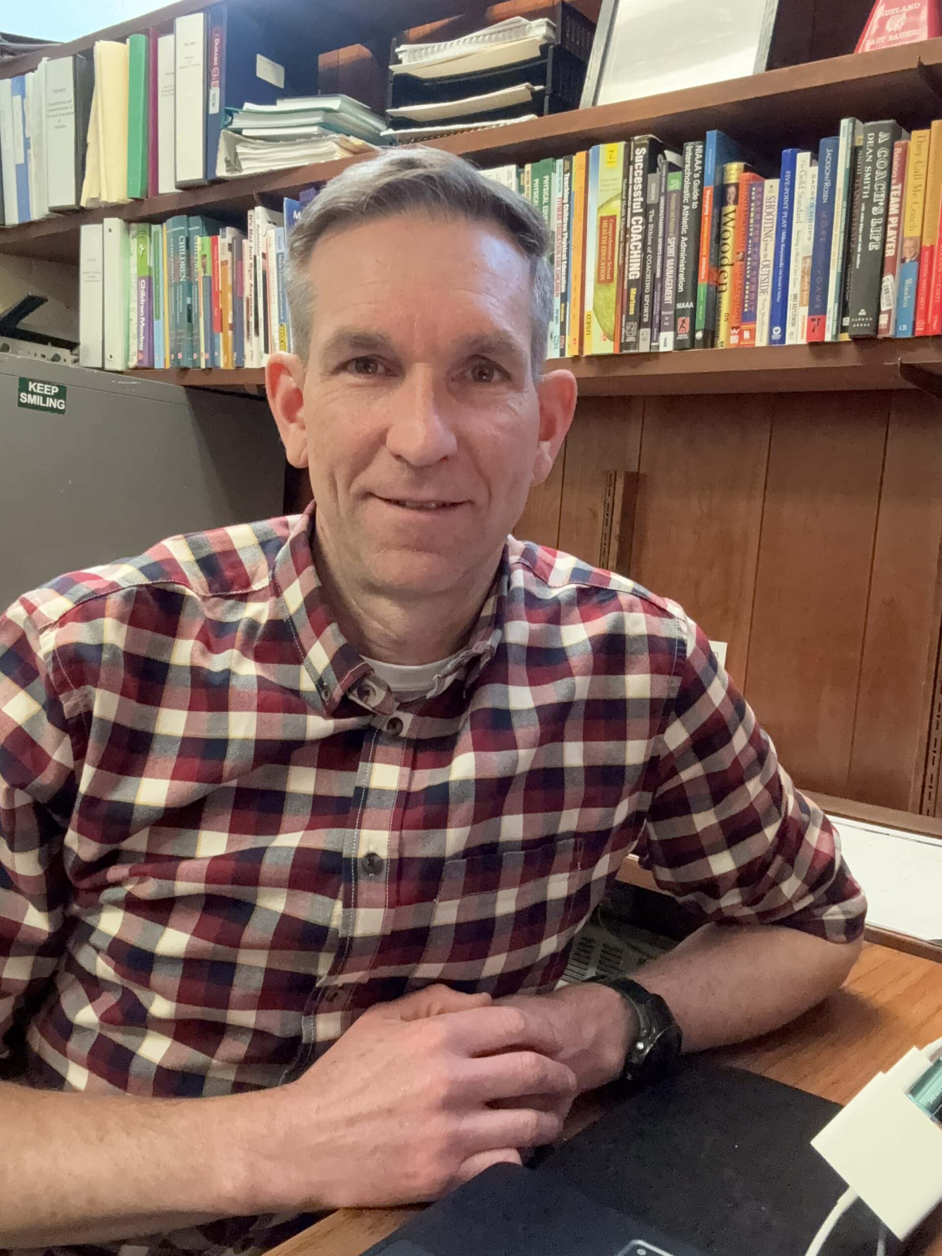 Tom Geisler wearing a plaid patterned shirt and smiling at his office desk