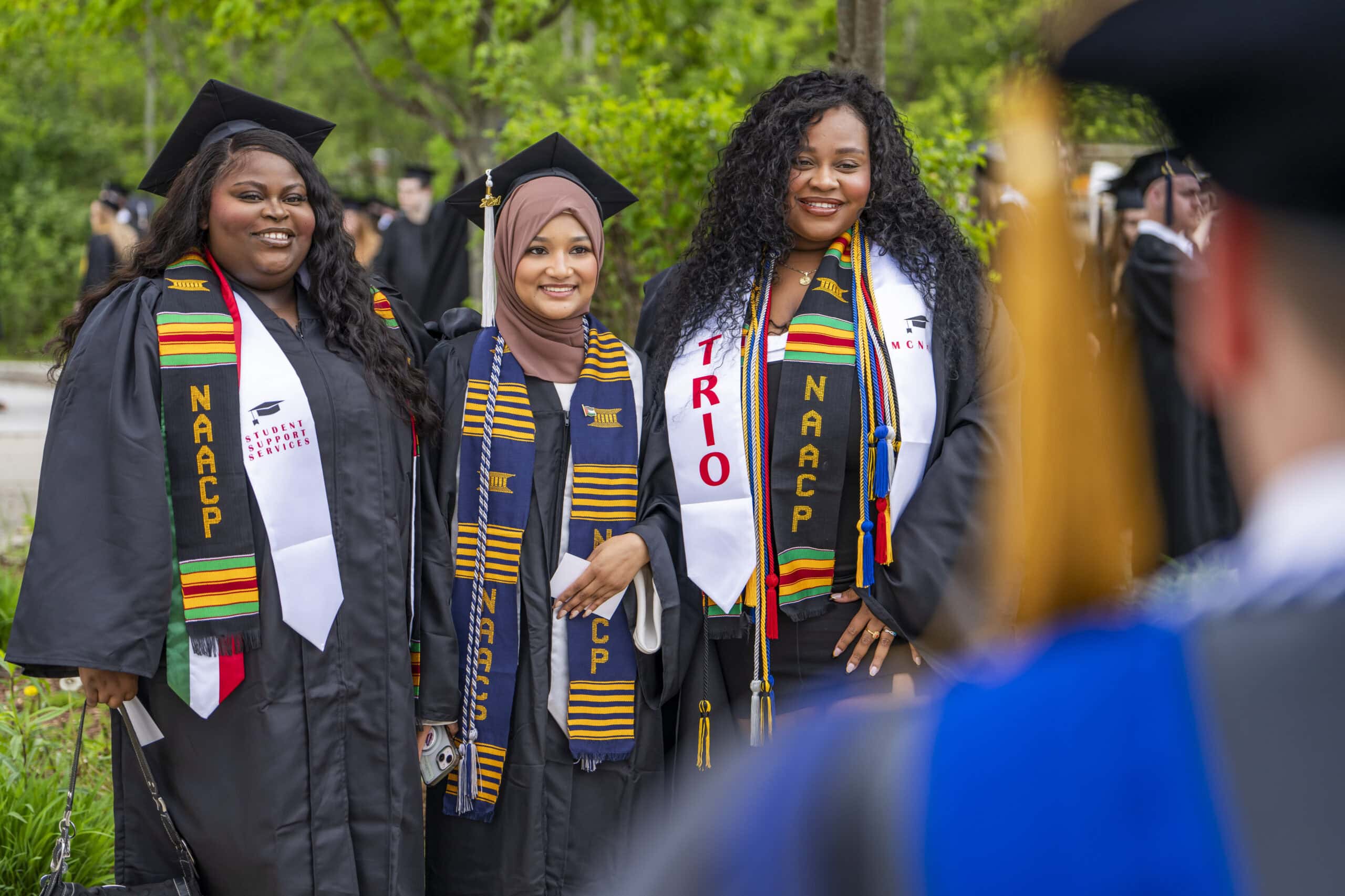 Three students of color wearing graduation gowns adorned with NAACP and TRIO stoles smiling