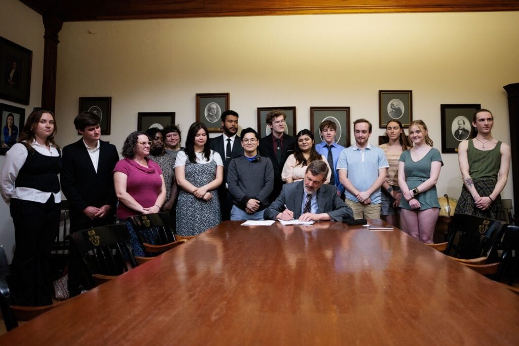 A group of student government leaders standing in a crowd behind VTSU President as he signs the common SGA constitution into action.