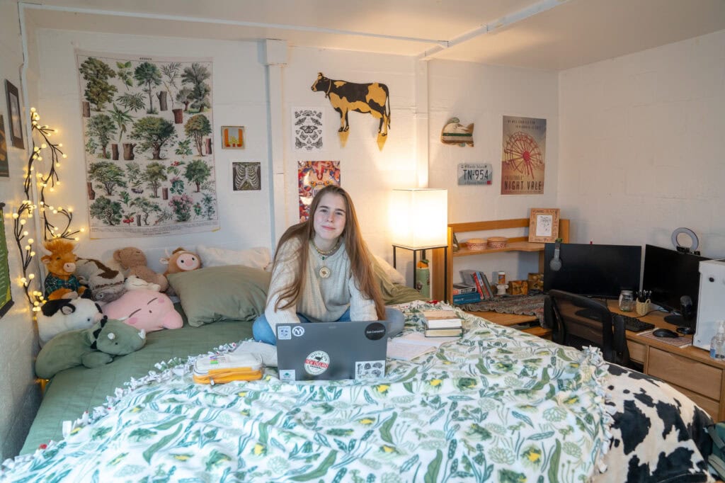 A young woman with long blonde hair sitting on her bed in a dorm at VTSU Castleton.