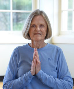 Faye Tolar smiling at the camera and holding her palms together in front of her body while wearing a blue shirt