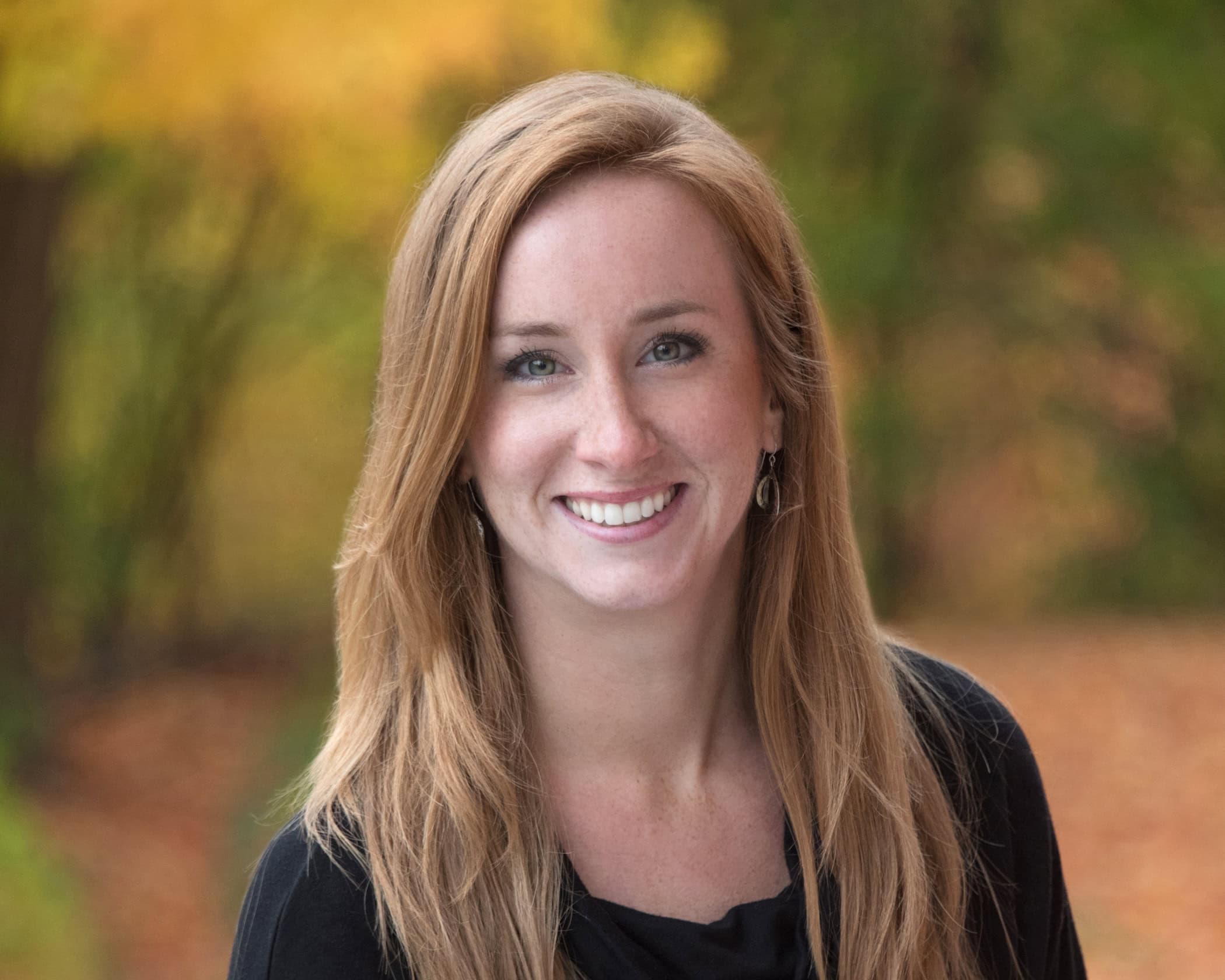 Kelly Dunklow smiling at the camera while wearing a black long sleeve shirt. She has long blonde hair and is standing outdoors on an autumn day with colorful foliage around her