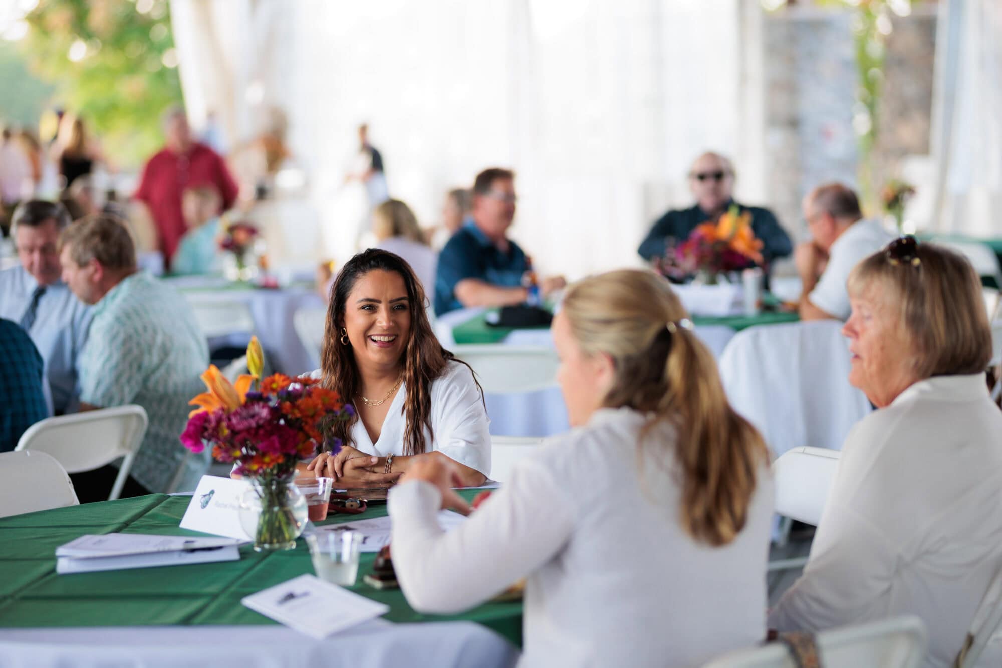 A group of people sitting at round tables decorated with white and green linens. Floral centerpieces are on every table, and three women dressed in white are sitting in the foreground