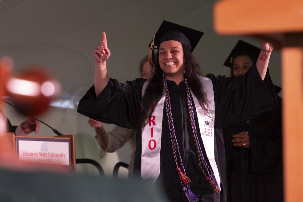 A graduate crossing the stage with their fingers crossed and arms raised above their head as they approach to get their diploma.