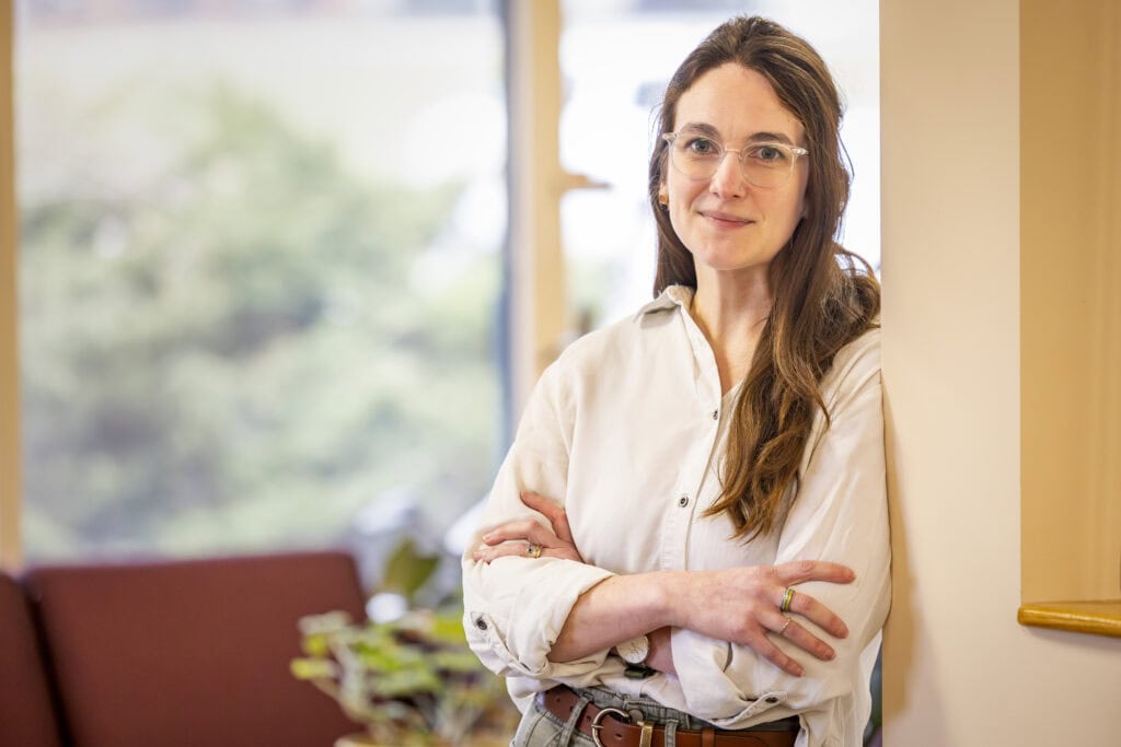 A woman stands leaning against a wall with her arms crossed in front of her, she is wearing glasses and there is a window behind her.