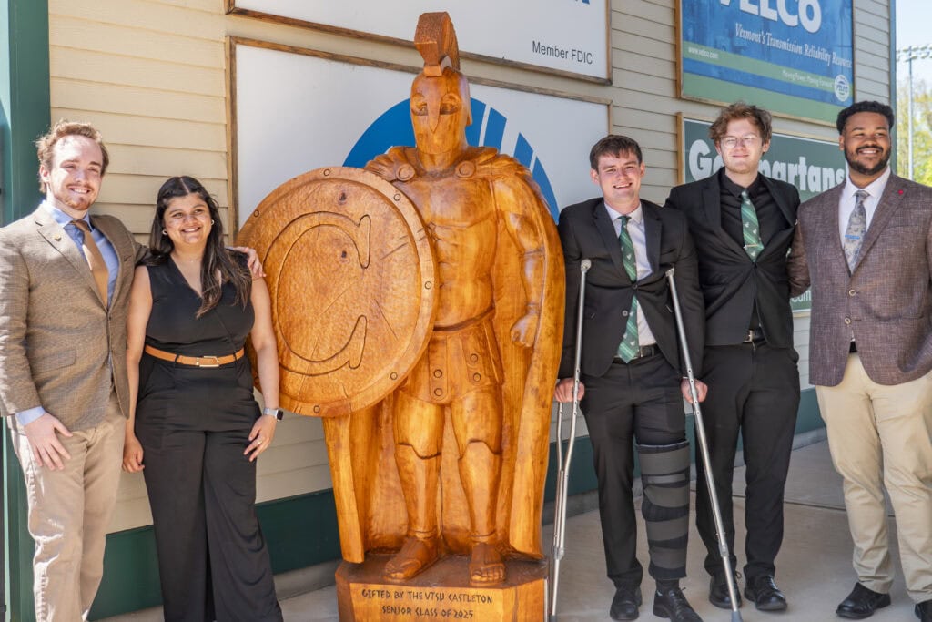 Students stand in group alongside carved wooden Spartan statue