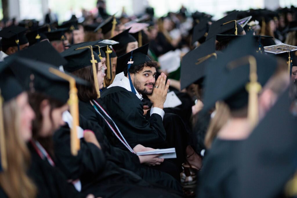 A large group of students in graduation caps and robes, a young man sitting in the middle of the crowd has his head turned toward the camera.