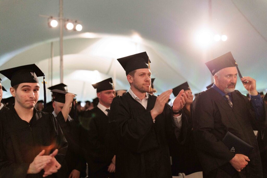 Three men in graduation caps and gowns sitting at the front of a crowd under a white event tent. 