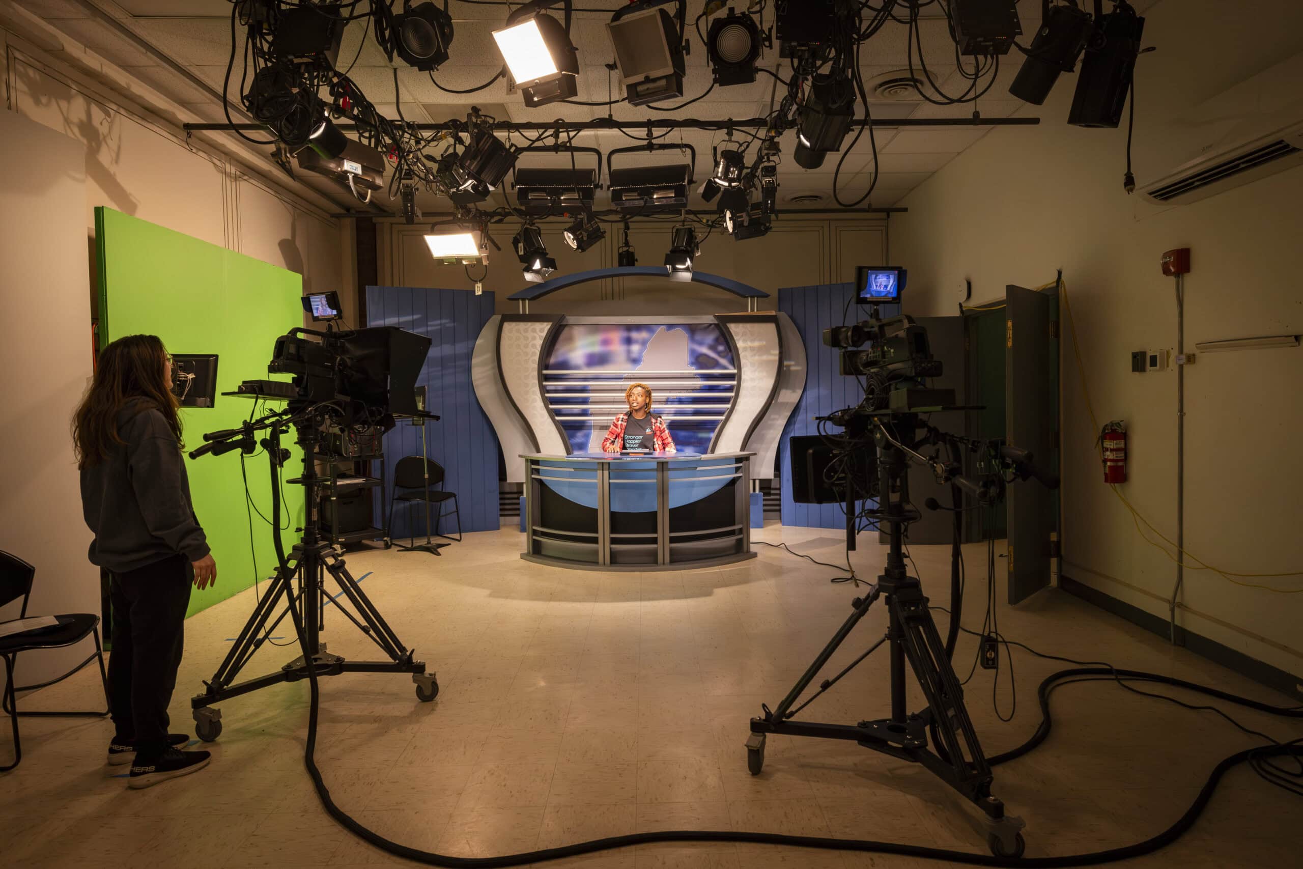 Two students working in the News7 Broadcast studio. One student is sitting behind the news desk while another stands behind a broadcast camera.