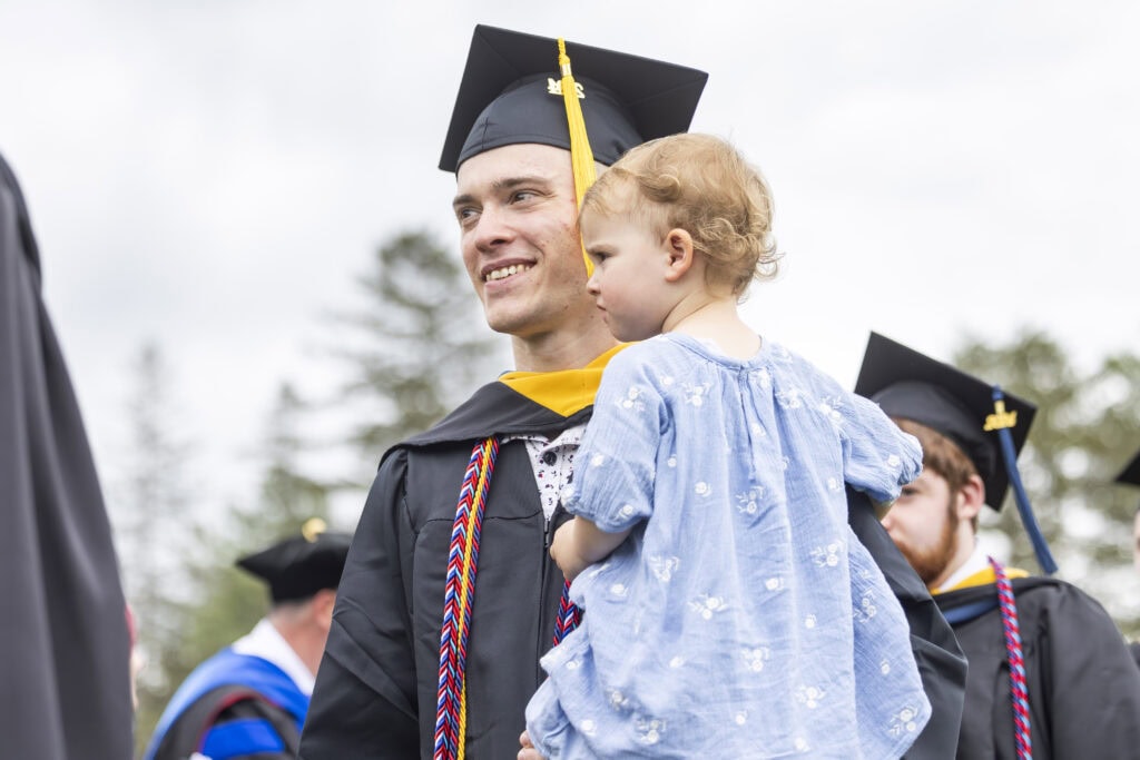 A man in graduation cap and gown holding his young daughter in his arms.