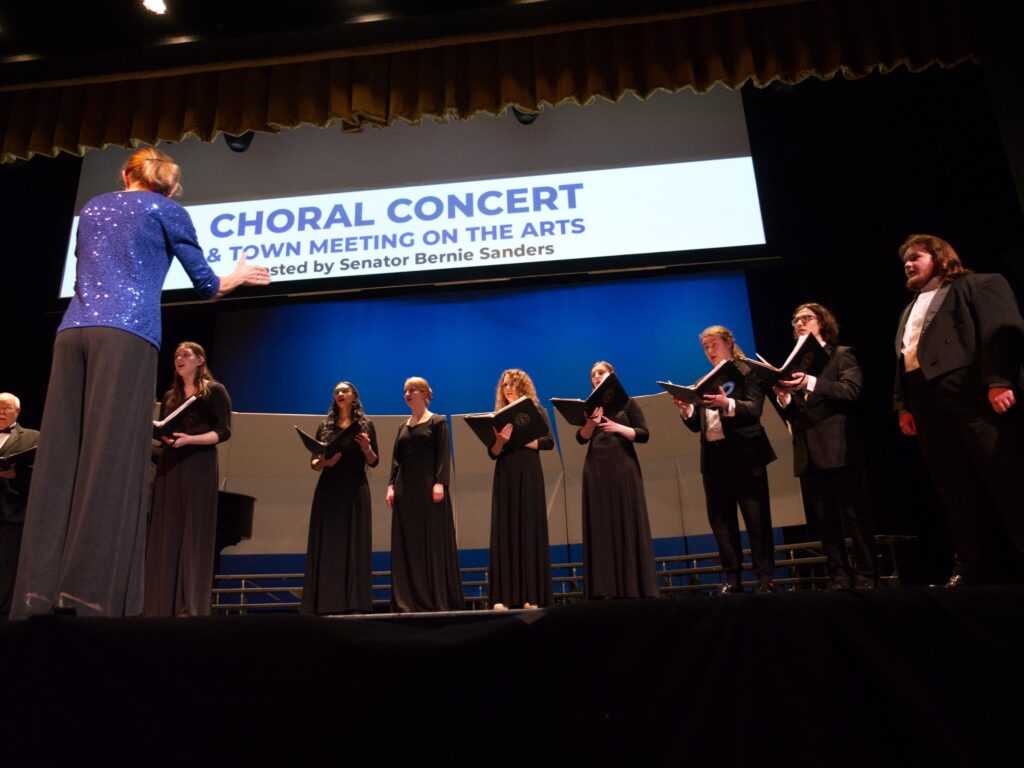 Members of the VTSU chamber singers performing on stage while Dr. Sherrill Blodget conducts. Some members wear long black gowns, and others wear black suits. All singers are holding a black folder of sheet music in their hands.