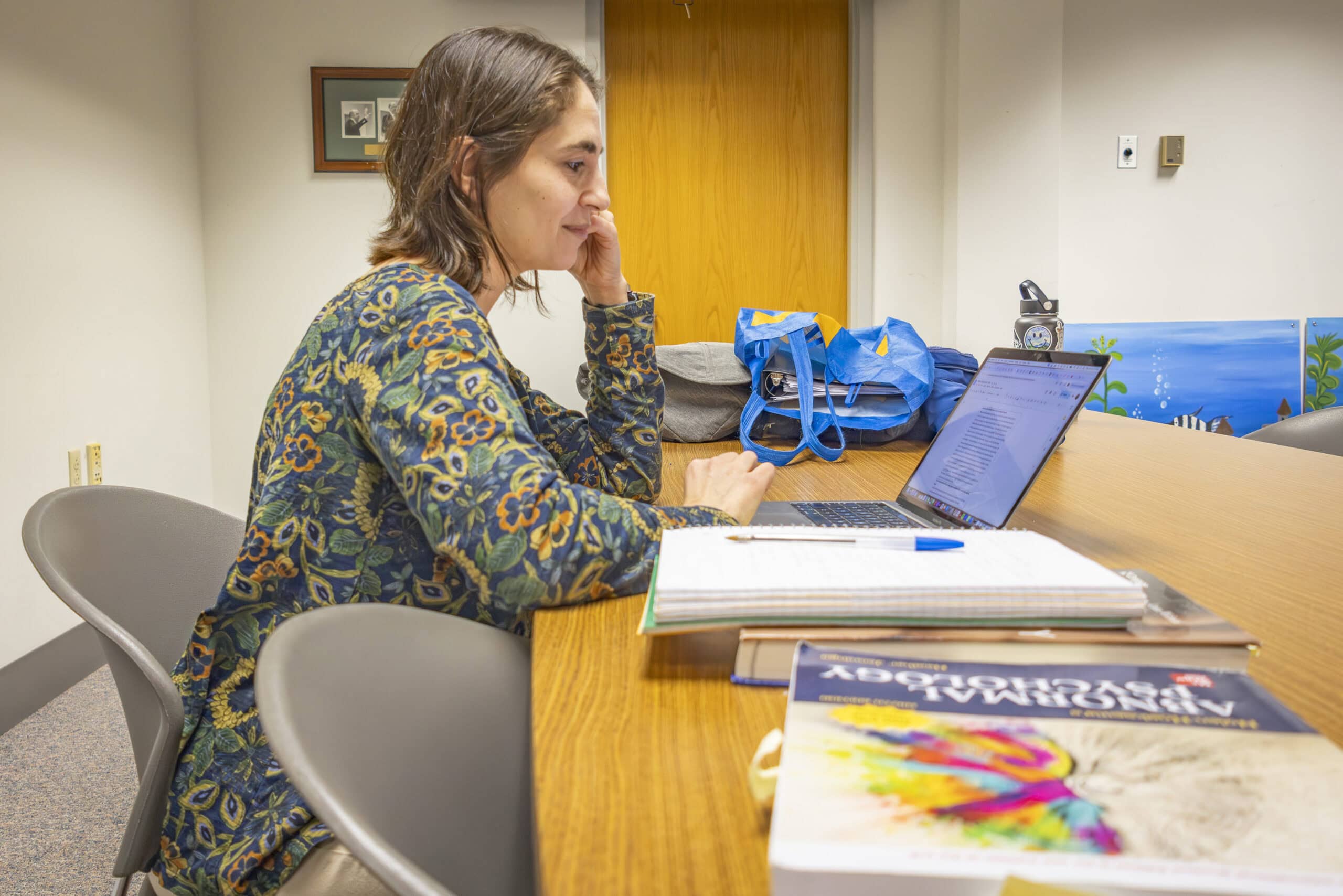 A woman wearing a floral patterned dress sitting at a long table. In front of her is a laptop, several textbooks, and a notebook with a pen balanced on top.