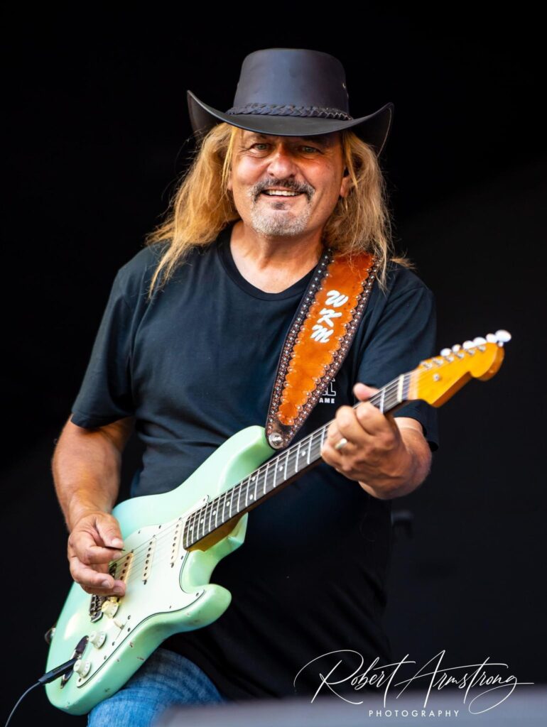 Kenny Mades, a man with long blonde hair wearing a cowboy hat and black t-shirt holding a mint blue electric guitar.