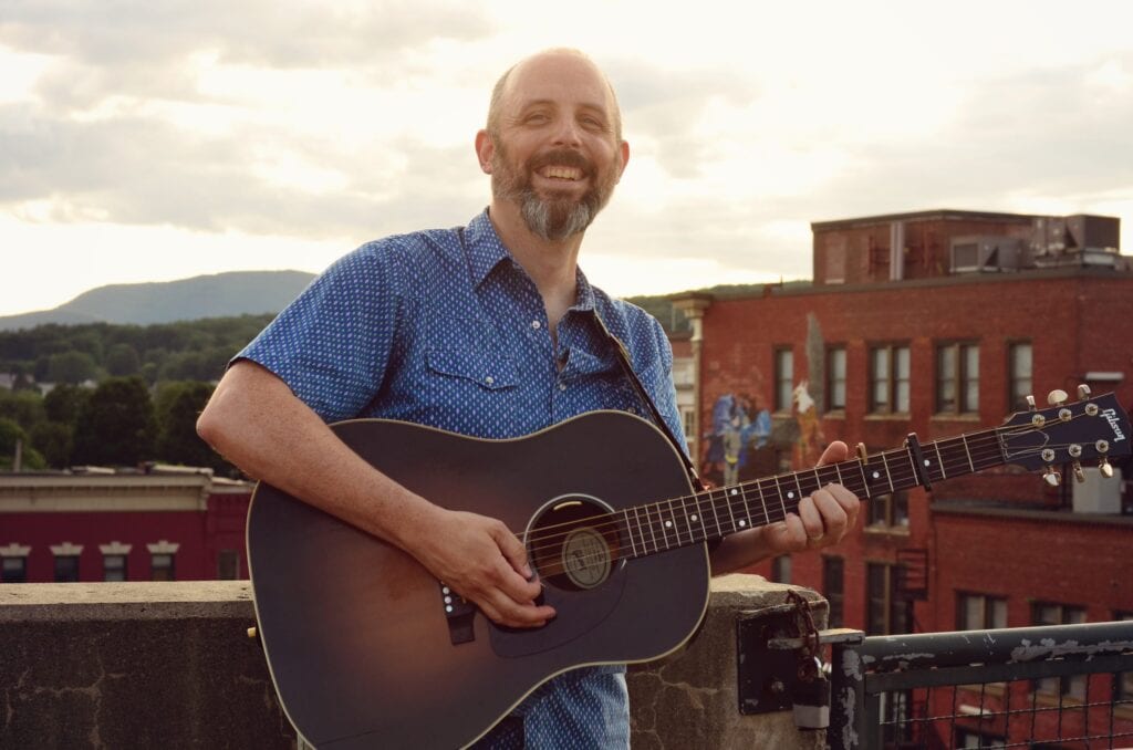 Phil Henry standing on a rooftop wearing a blue polo shirt and holding a dark brown acoustic guitar.