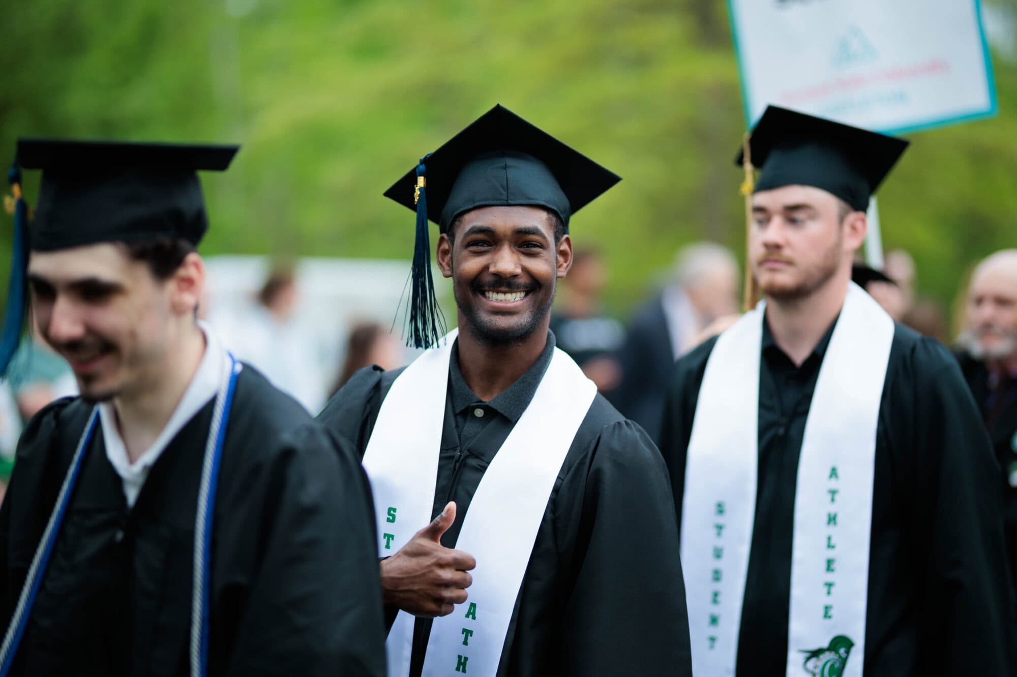Men in graduation regalia stand in a line, and the man in the center smiles and gives a thumbs up.