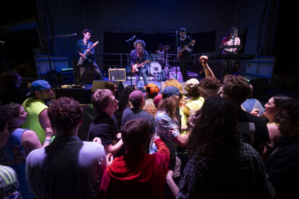 A three-member band performing on the outdoor stage at Greggfest on the Lyndon campus. A large group of students is gathered at the front of the stage.