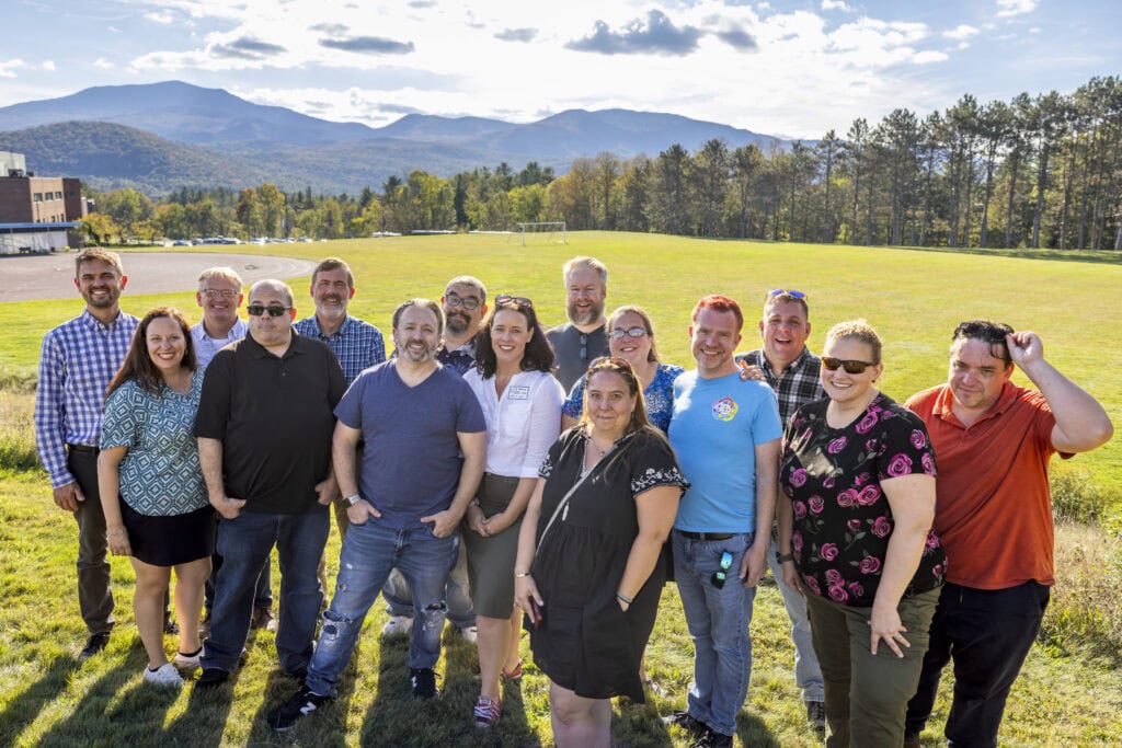 A large group of alumni standing together and smiling at the camera. They're standing in a large grassy field with mountains and blue sky in the background.