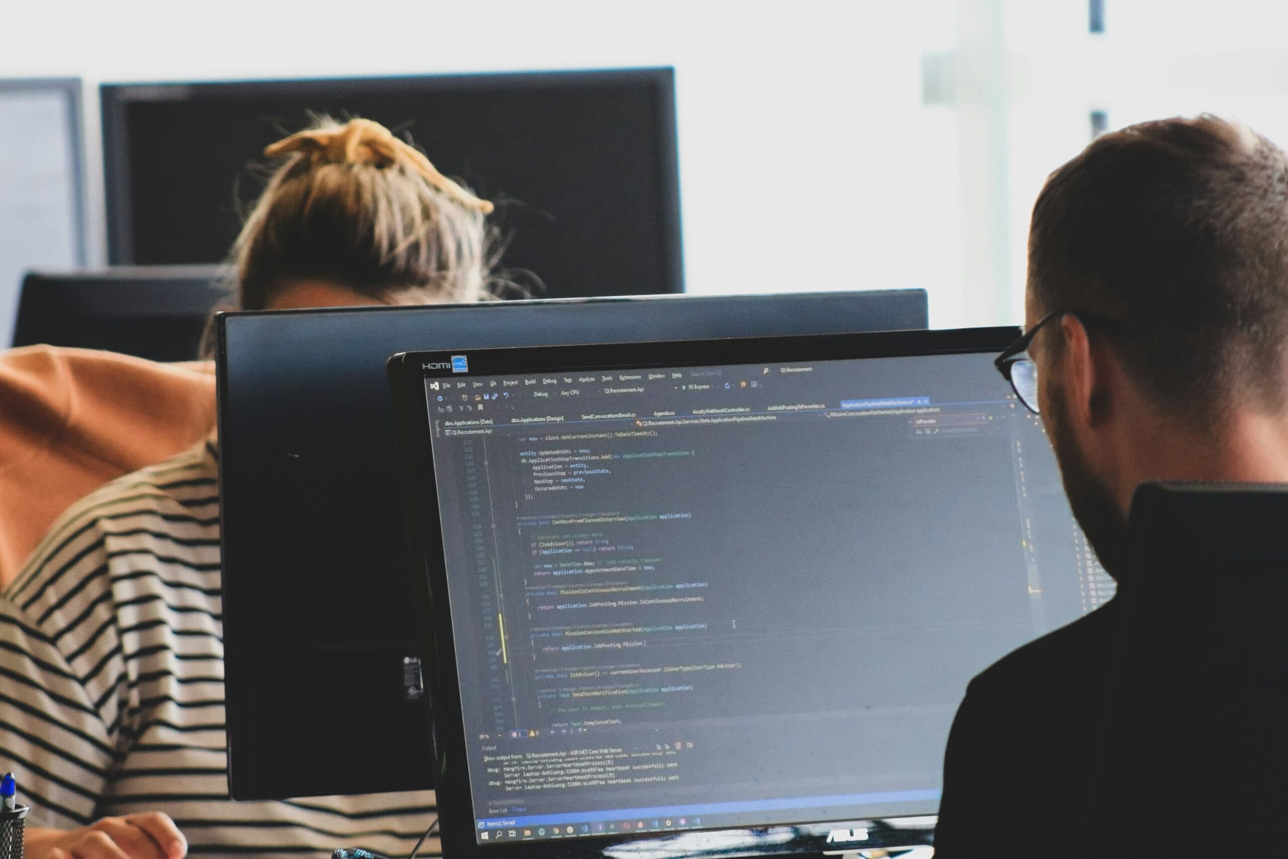 A man sitting in front of a computer while programming, with his back to the camera. A woman with blonde hair is sitting at a computer opposite him, her face obstructed.
