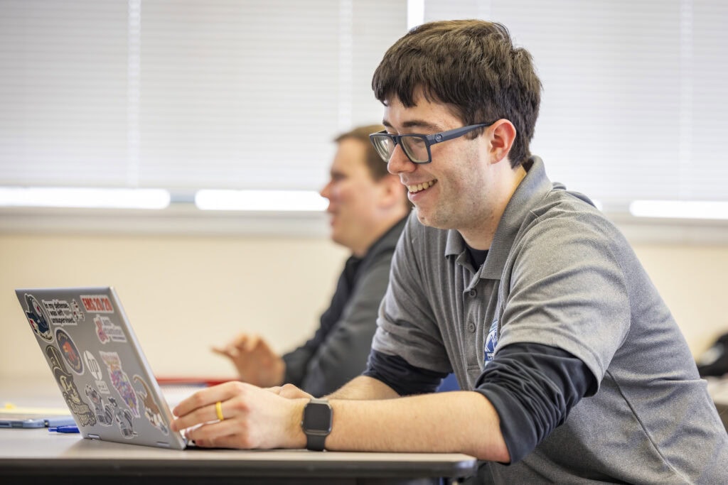A man with short curly brown hair sits in a classroom looking at a laptop.