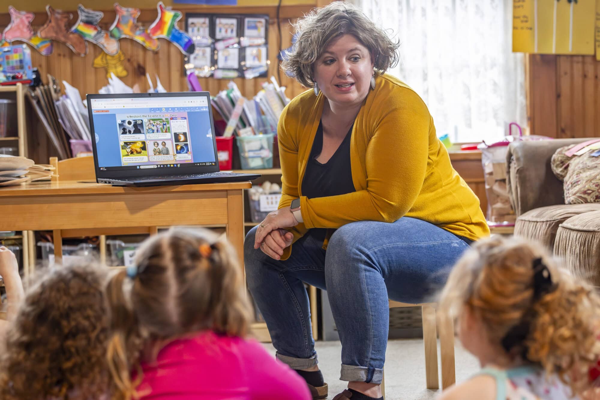 A teacher sits at a laptop in a classroom facing students, leaned forward with her elbows on her legs.